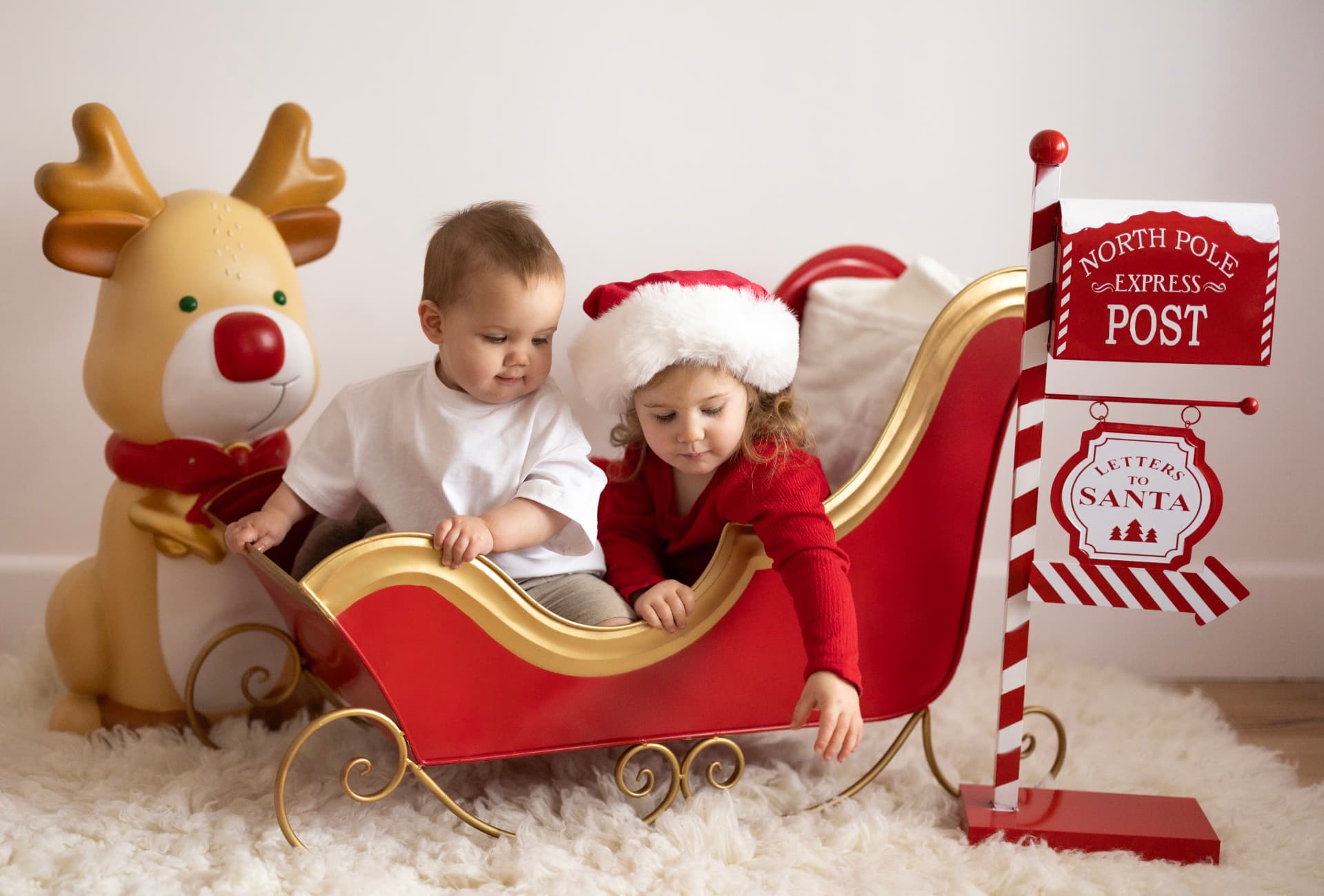 Two young children sitting in a red Christmas sleigh with a reindeer