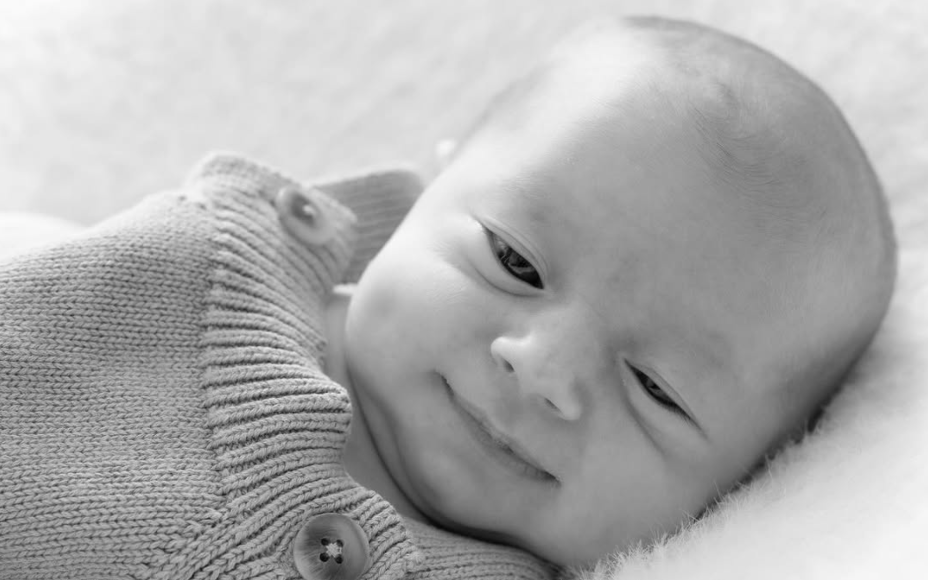 Newborn baby in a knitted cardigan resting on soft bedding, black and white
